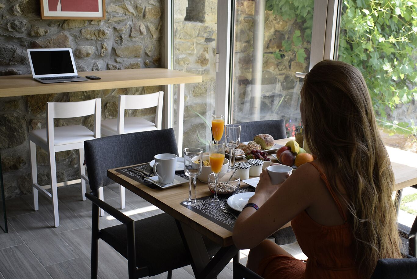 Une table mise pour le petit-déjeuner, avec un ordinateur portable, une tasse, un verre de jus d’orange et une assiette de nourriture.