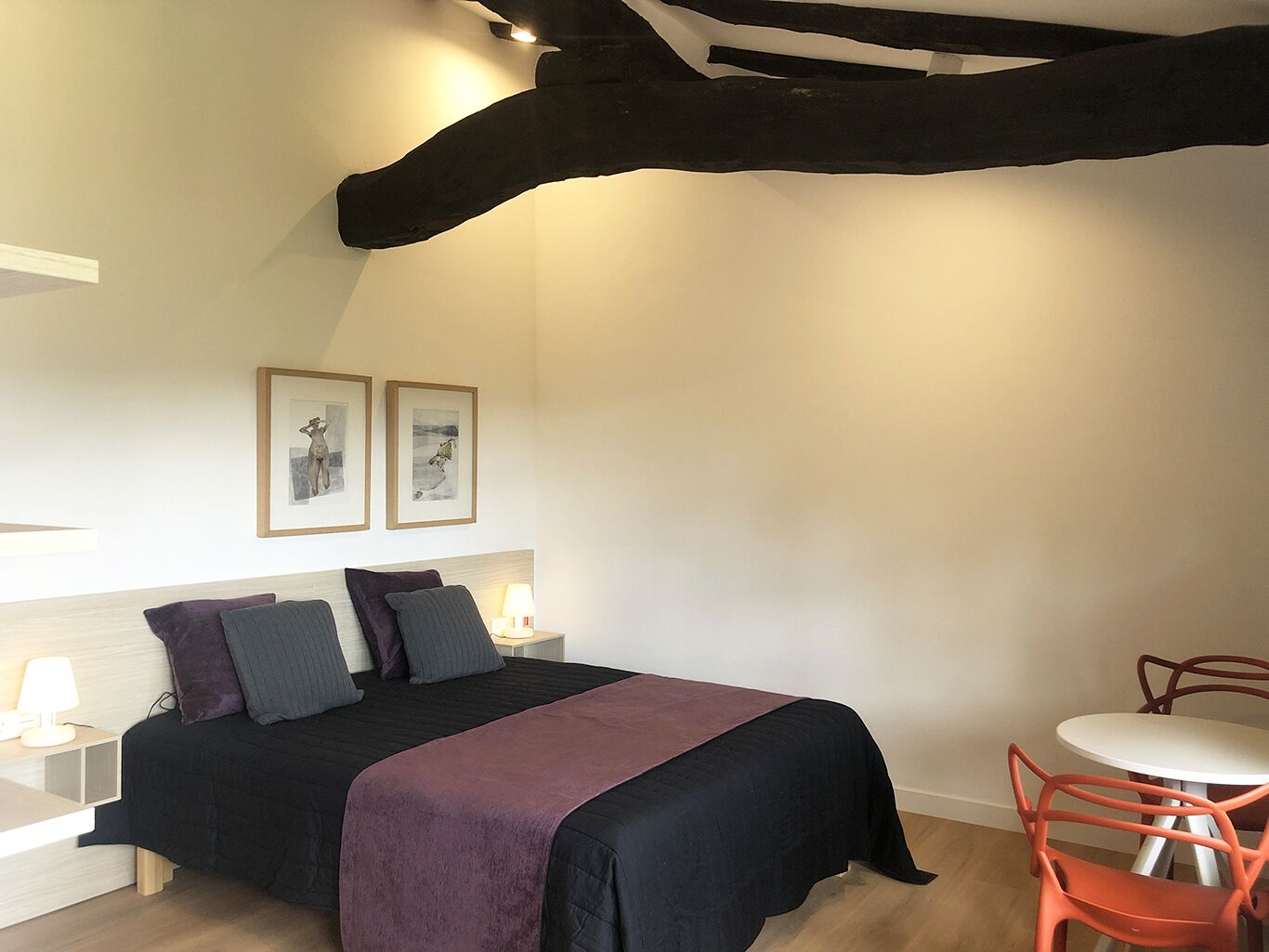 A bedroom with a bed, two pillows, a bedside table with a lamp, a small table with chairs, and two framed pictures on the wall.