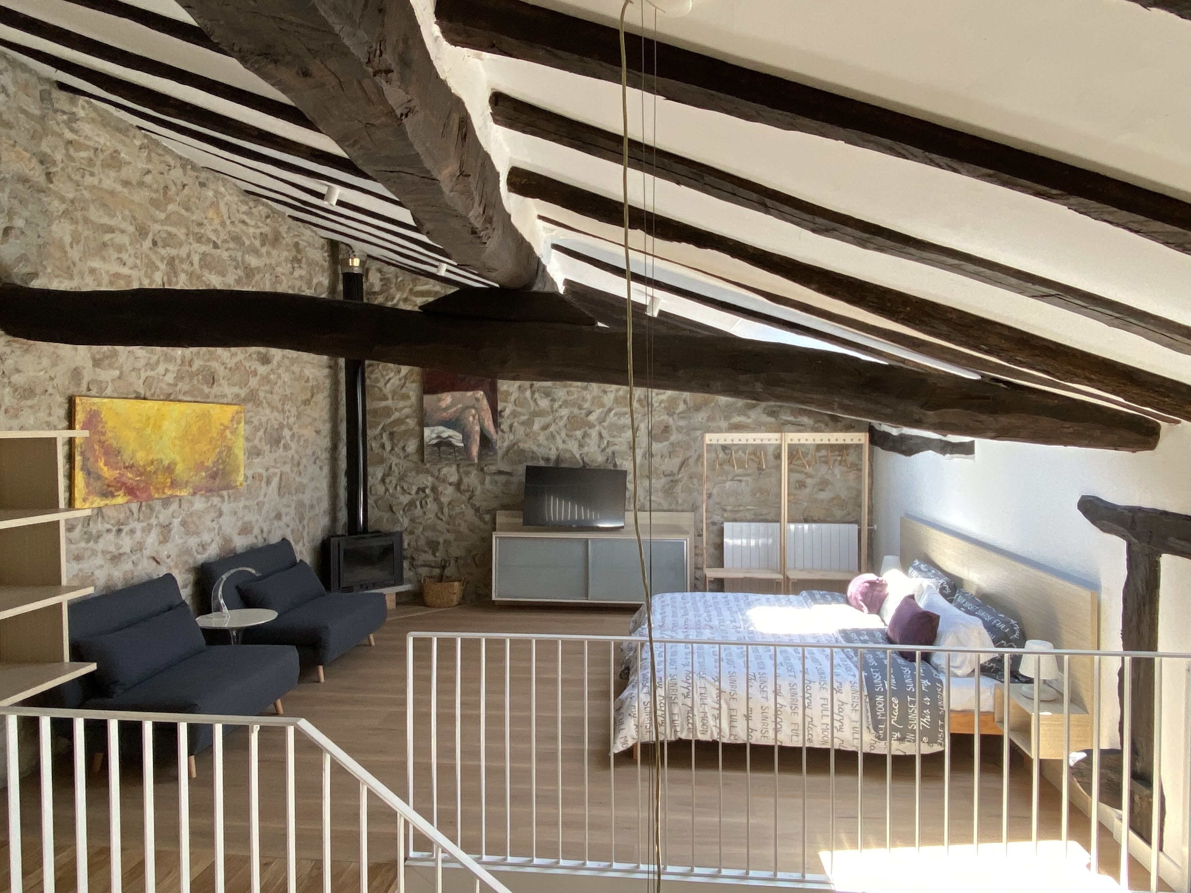 Une chambre à coucher au style rustique, avec des poutres apparentes, un mur en pierre, un lit avec une literie à motifs, une télévision et un petit coin salon.