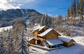 Una cabaña de madera con balcón y zona de estar al aire libre techada, rodeada de árboles cubiertos de nieve y una montaña al fondo.