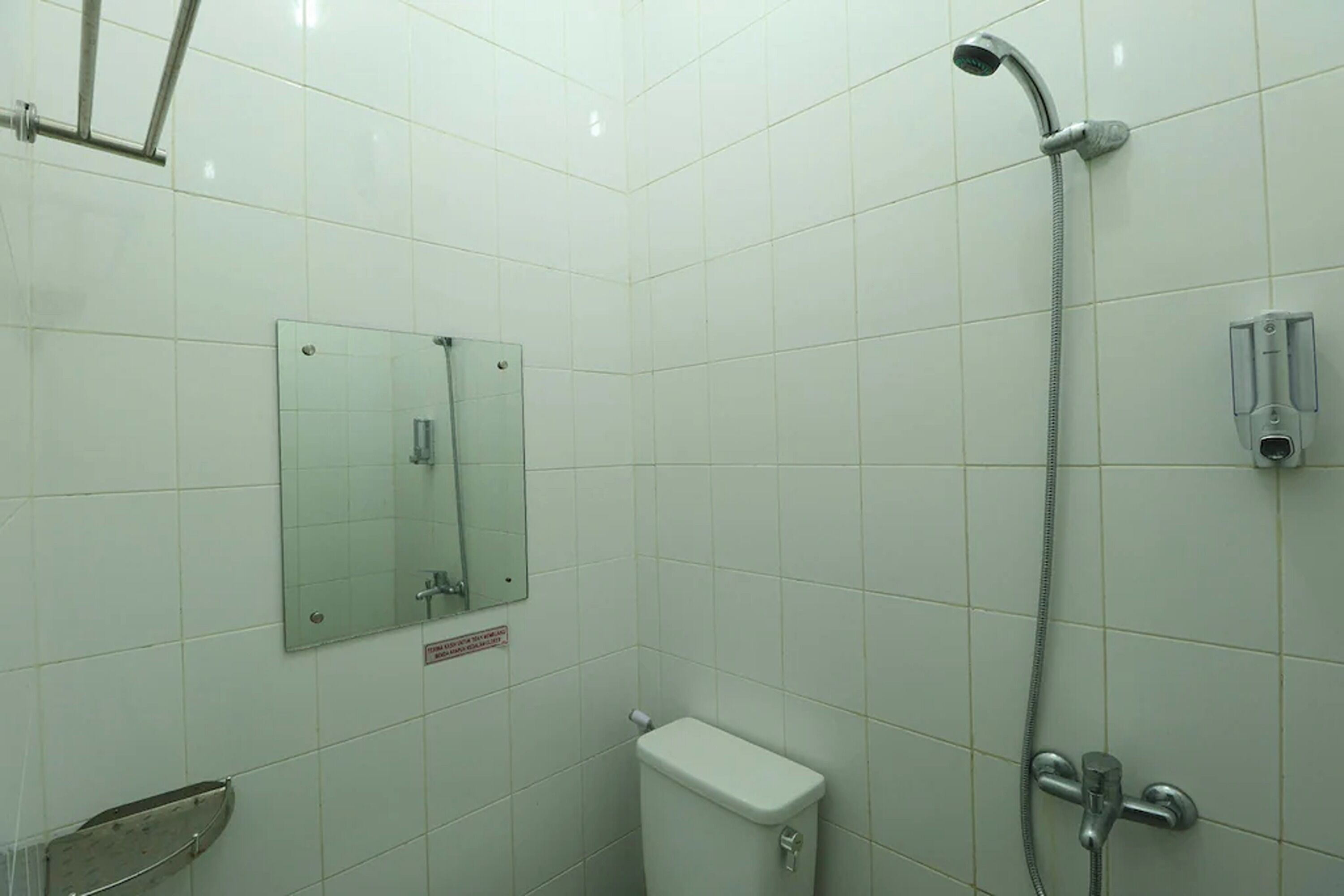 A bathroom with white tiles, a toilet, a shower with a handheld showerhead, and a mirror with a warning sign.