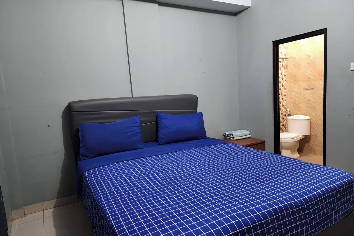 Quarto com cama, duas almofadas, mesa de cabeceira e espelho refletindo um banheiro.