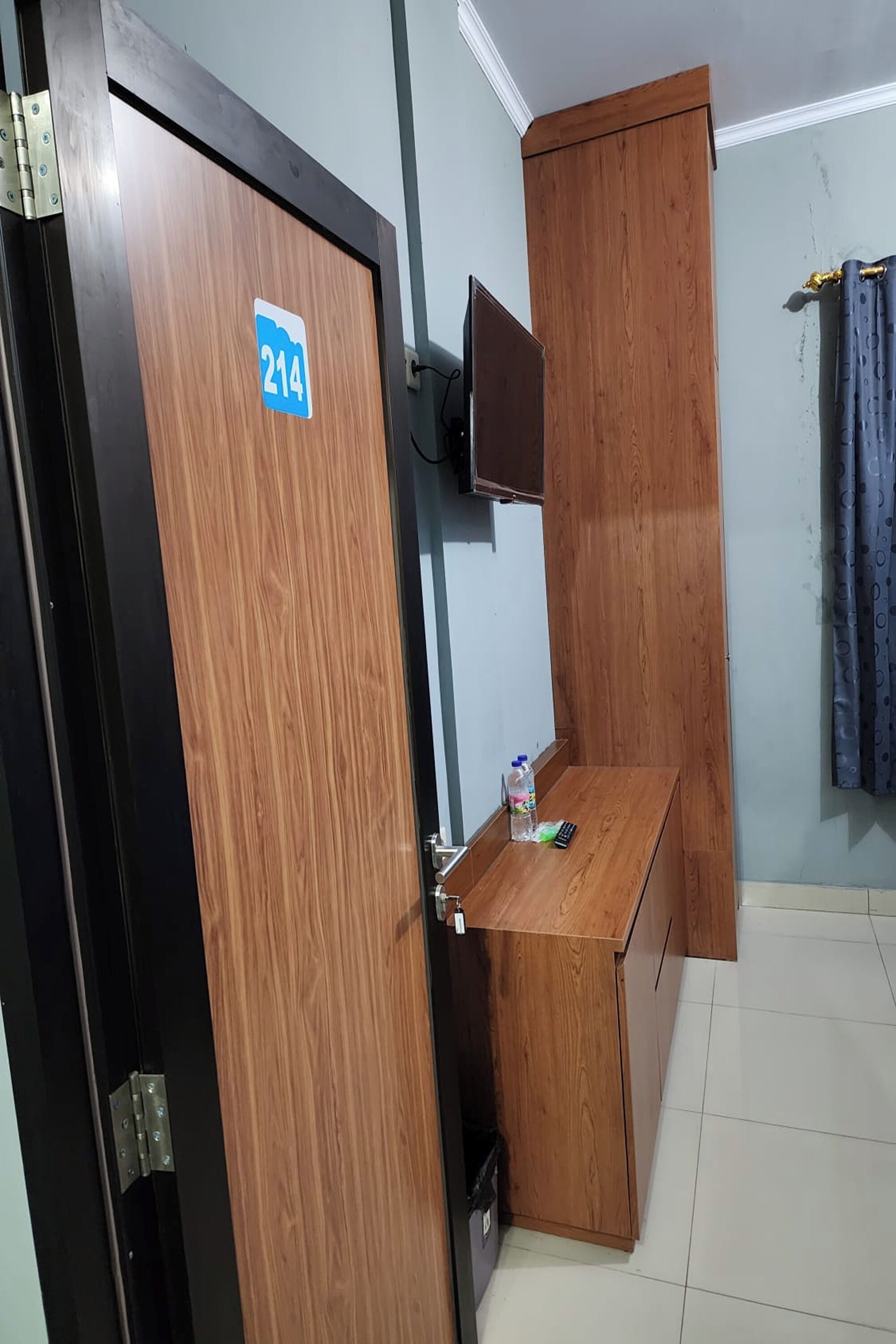 Una habitación con una puerta de madera que indica el número 214, un pequeño escritorio de madera con una botella y un control remoto, un televisor montado en la pared y una cortina en la ventana.