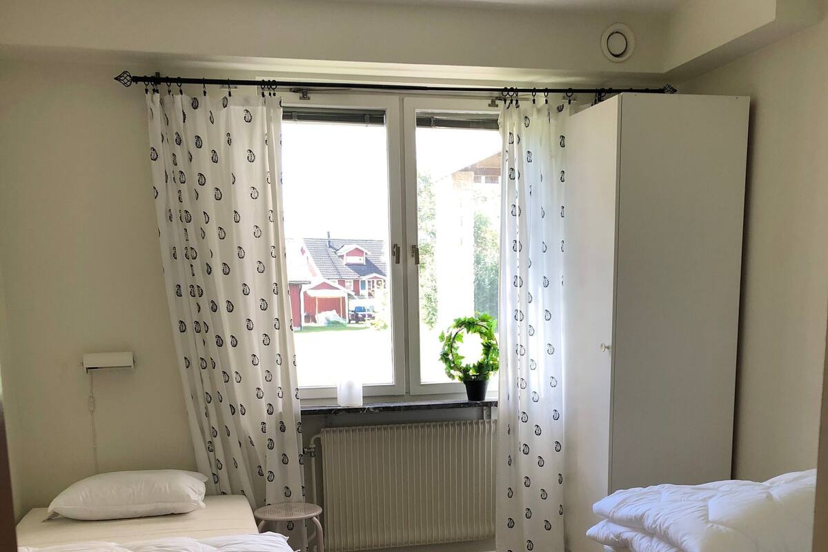 A bedroom with two beds, a radiator, a window with curtains, a small table, and a potted plant.