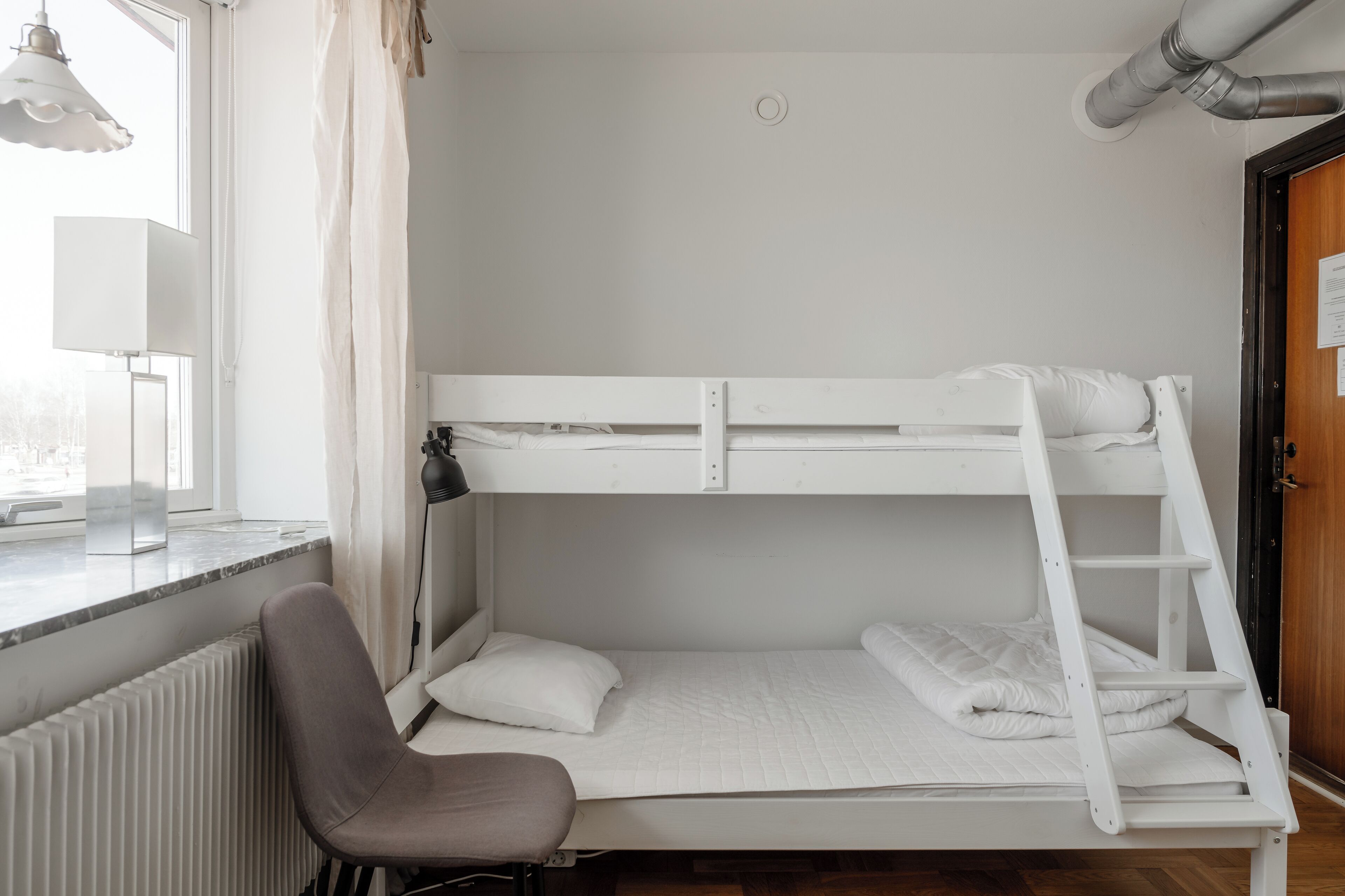 A bunk bed with a ladder, a chair, a radiator, and a window with curtains.