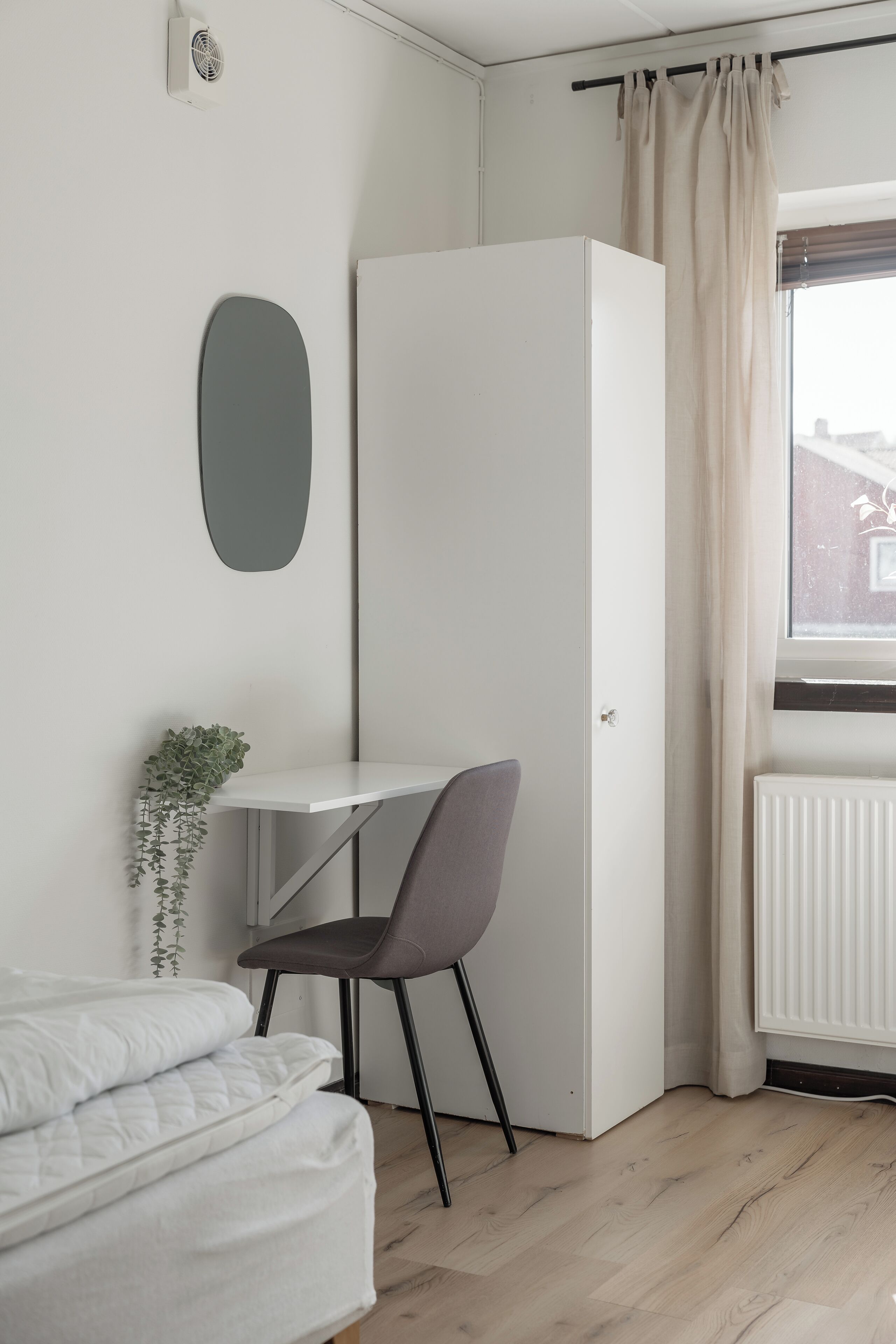 A compact bedroom with a bed, a small desk, a chair, a radiator, and a window with curtains.