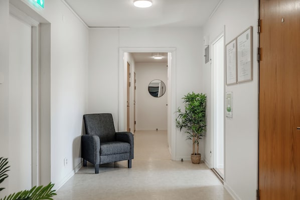 A hallway with a grey armchair, a potted plant, and framed documents on the wall.