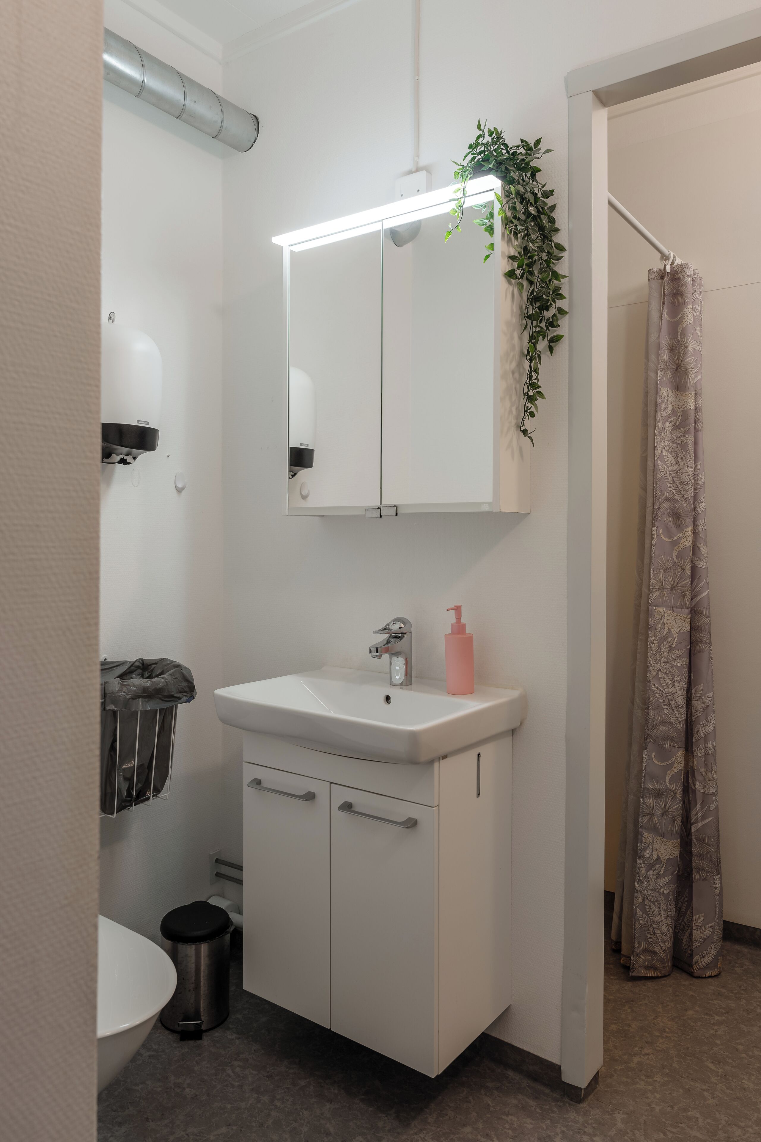 A bathroom with a white sink, mirror, and cabinets, a trash can, and a shower curtain.