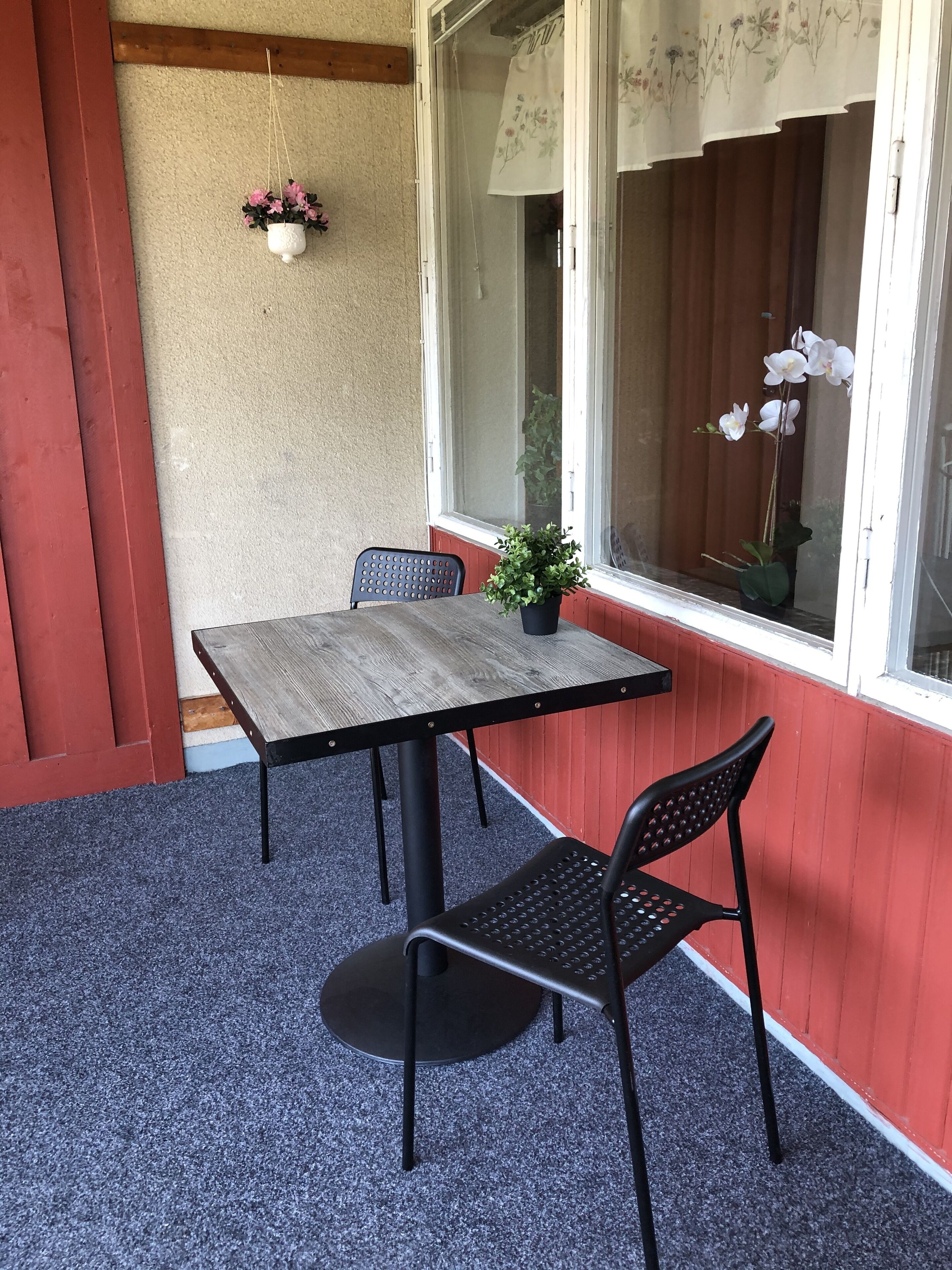A small patio with a wooden table and two chairs, a hanging flower pot, and a window with curtains.