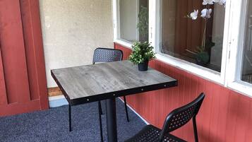 Un pequeño patio con una mesa de madera y dos sillas, una maceta colgante y una ventana con cortinas.