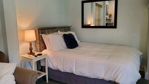 A neatly made bed with white linens, a small bedside table with a lamp, and a chair with a cushion. - Studio - direct 2 min beach walk with public access end of street. (Oak Island)