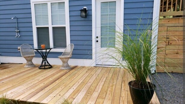 A wooden deck with two wicker chairs and a table, a potted plant, and a house with a blue exterior and white doors. - Studio - direct 2 min beach walk with public access end of street. (Oak Island)