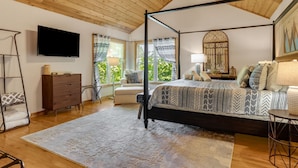 A four-poster bed with a patterned bedspread, a television on the wall, and a view of greenery through large windows.