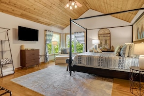 A four-poster bed with a patterned bedspread, a television on the wall, and a view of greenery through large windows.