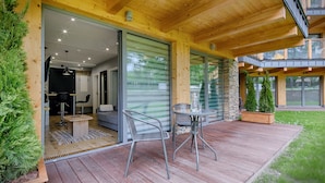 A wooden deck with a table and chairs, a stone wall, and a glass door leading to a room with a sofa and a dining table. - Goralski Szyk Royal Resort Wellness&SPA (Zakopane)