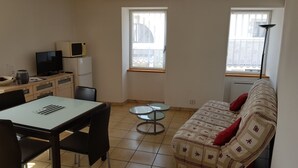 Un espacio habitable compacto con una mesa de comedor, sillas, un sofá, una pequeña cocina con microondas y refrigerador, y dos ventanas que aportan luz natural.