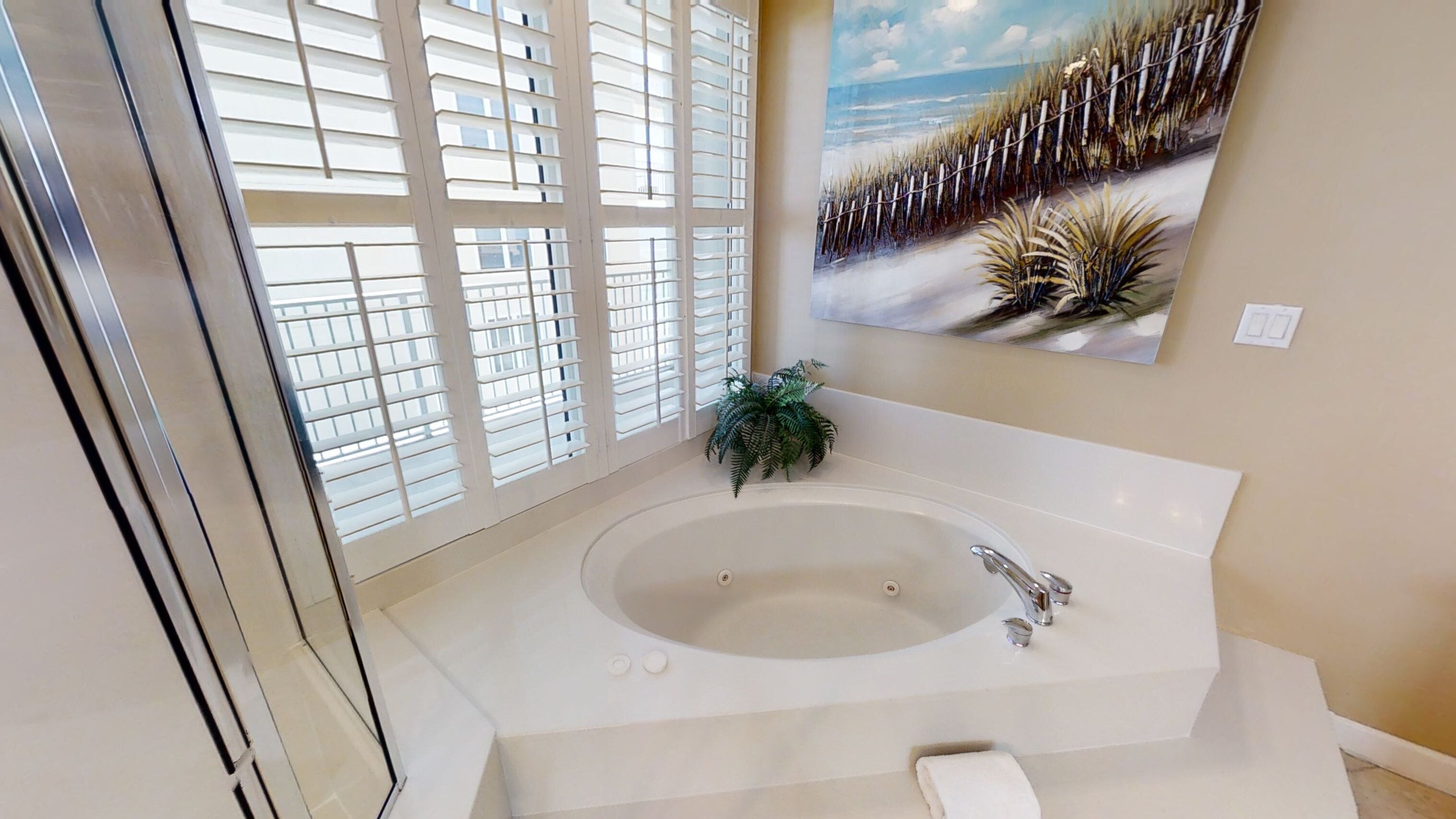 A bathroom with a large bathtub, a potted plant, and a beach-themed painting on the wall.