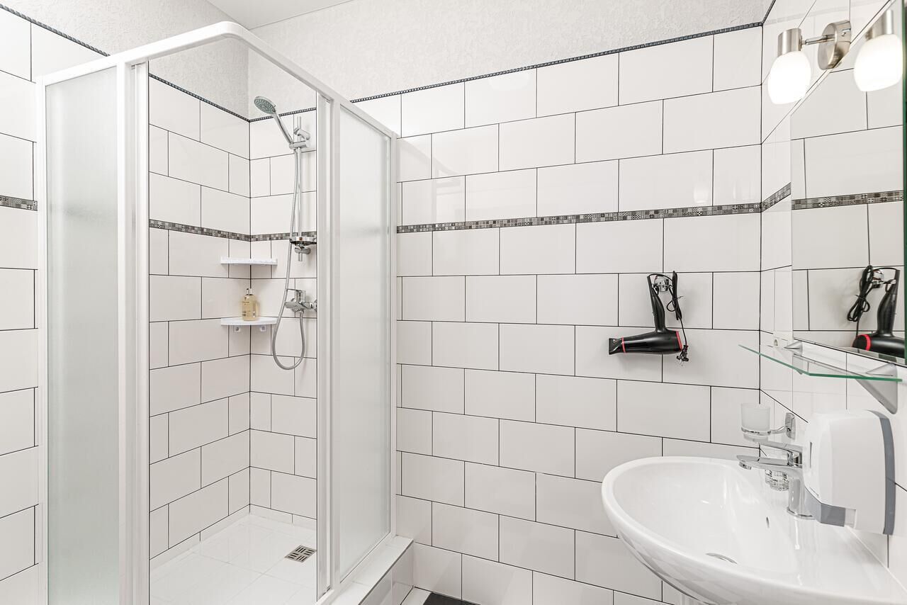 A modern bathroom with white tiles, a glass shower enclosure, a white sink, and a mirror with a glass shelf.