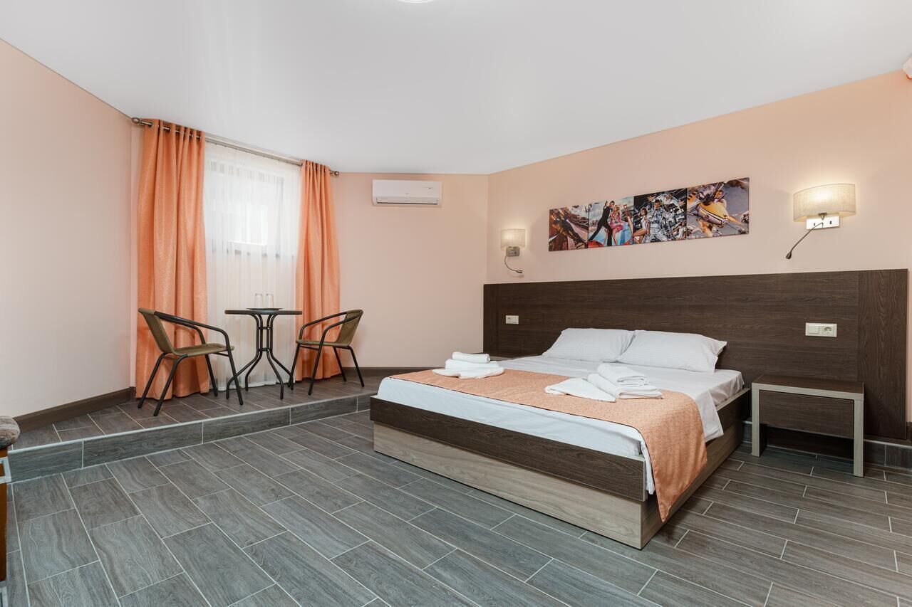 A hotel room with a bed, a small table, two chairs, an air conditioning unit, a wall-mounted lamp, and a painting.
