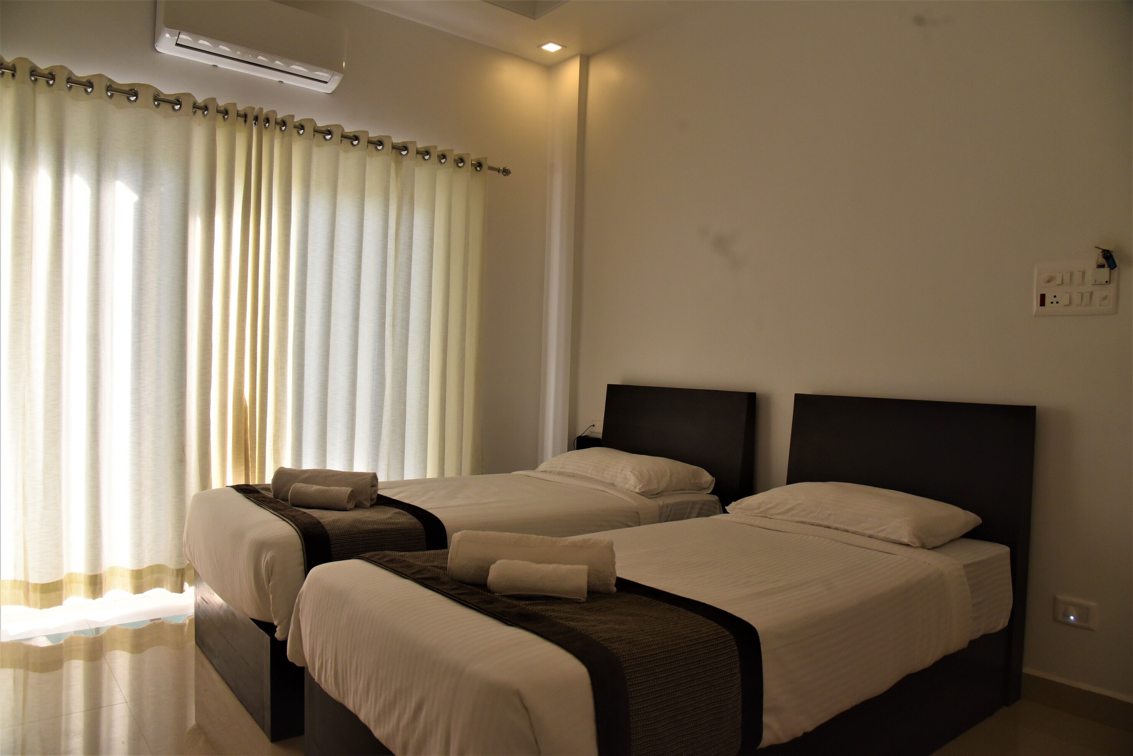 A hotel room with two beds, a large window with curtains, an air conditioning unit, and a wall socket.