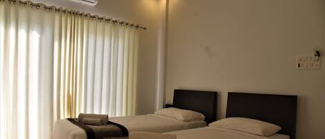 A hotel room with two beds, a large window with curtains, an air conditioning unit, and a wall socket.