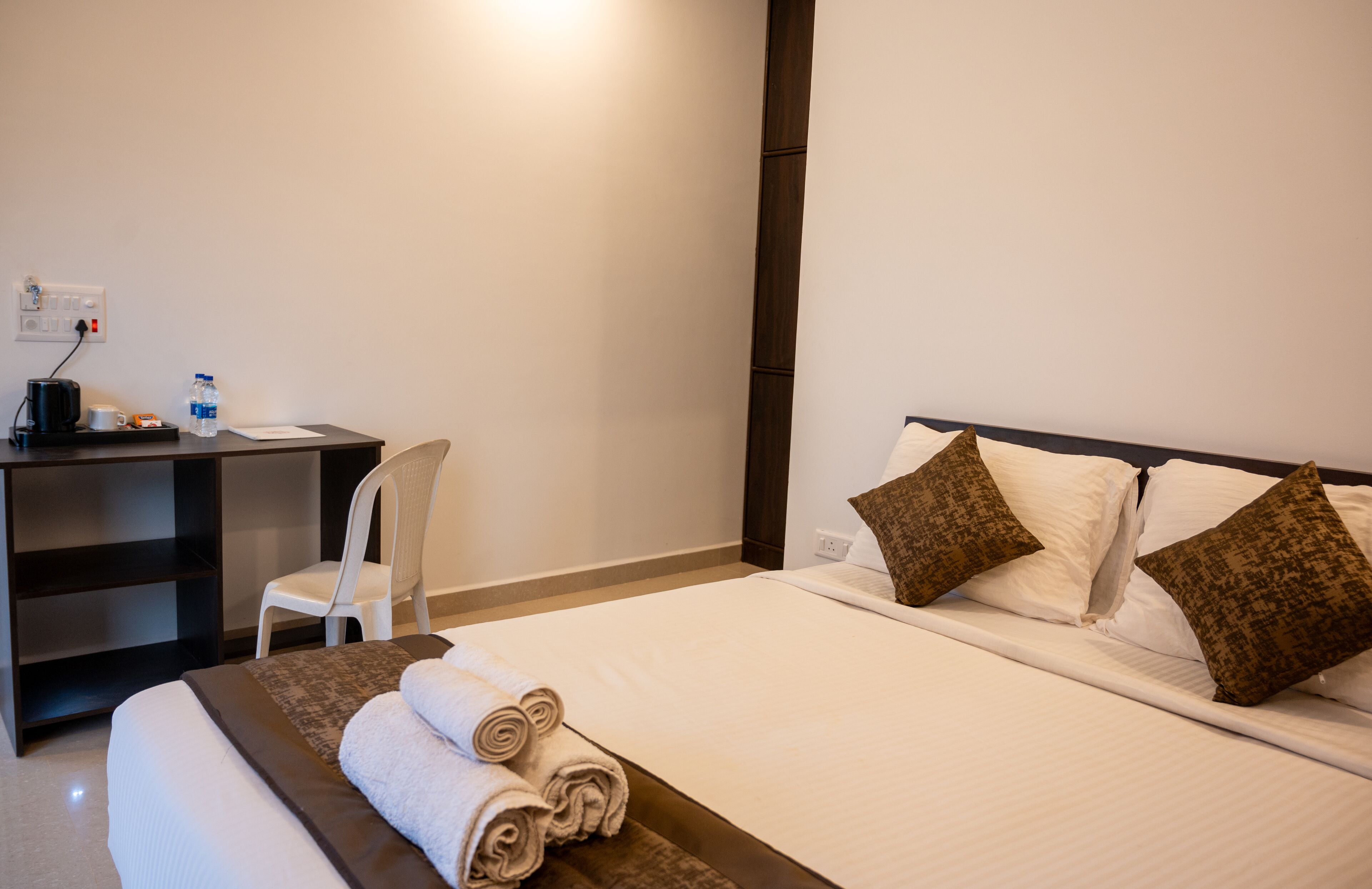 A hotel room with a neatly made bed, a desk with a chair, and a small table with a water bottle and a cup.