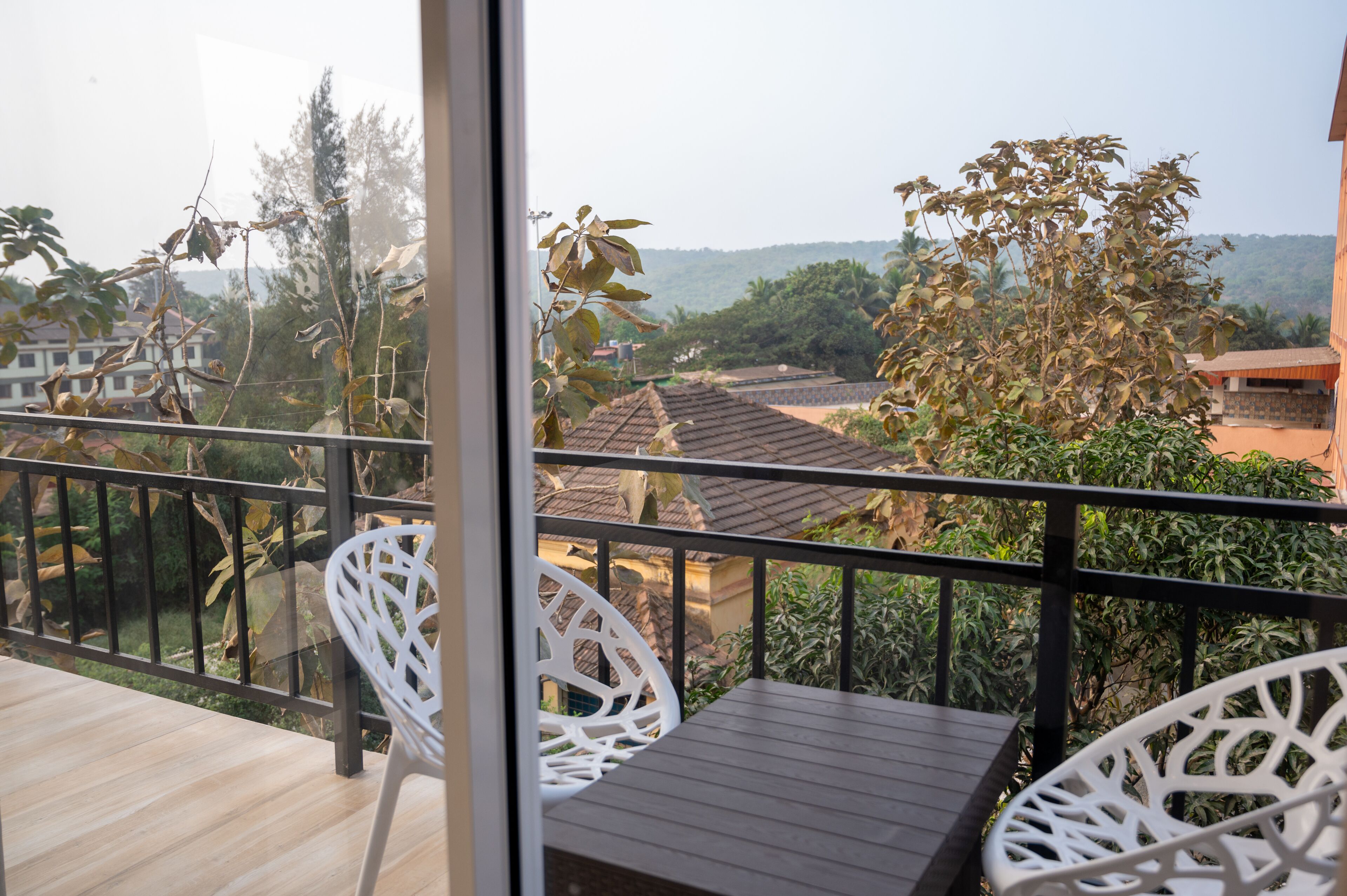 Een balkon met witte rieten stoelen en een houten tafel, met uitzicht op een woonwijk met bomen en huizen.