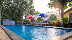 A swimming pool with lounge chairs and umbrellas, surrounded by trees and a building with a sign. - Aaria Hills (Arambol)
