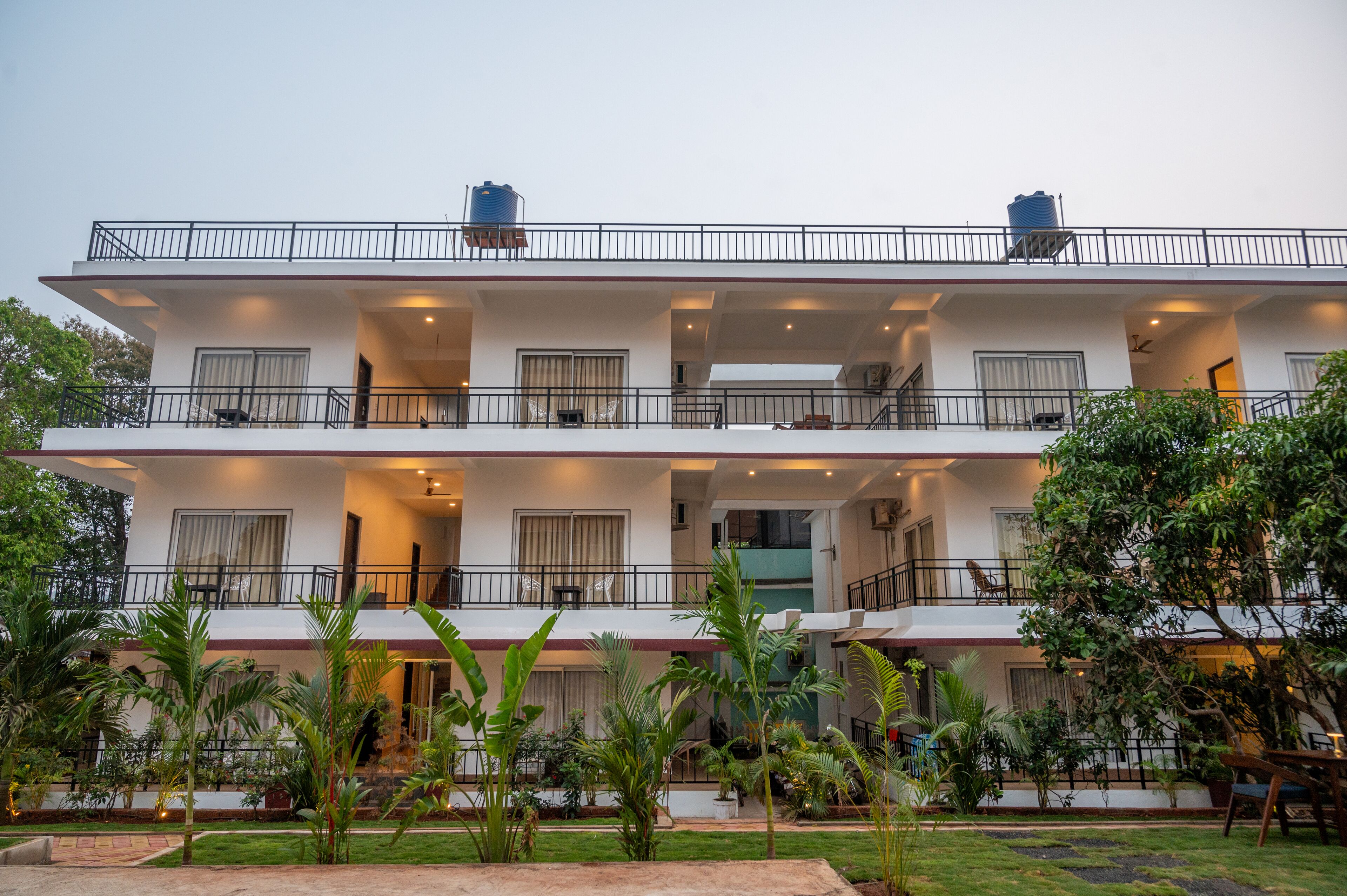 Un edificio de dos pisos con balcones, ventanas amplias y un jardín con plantas tropicales.