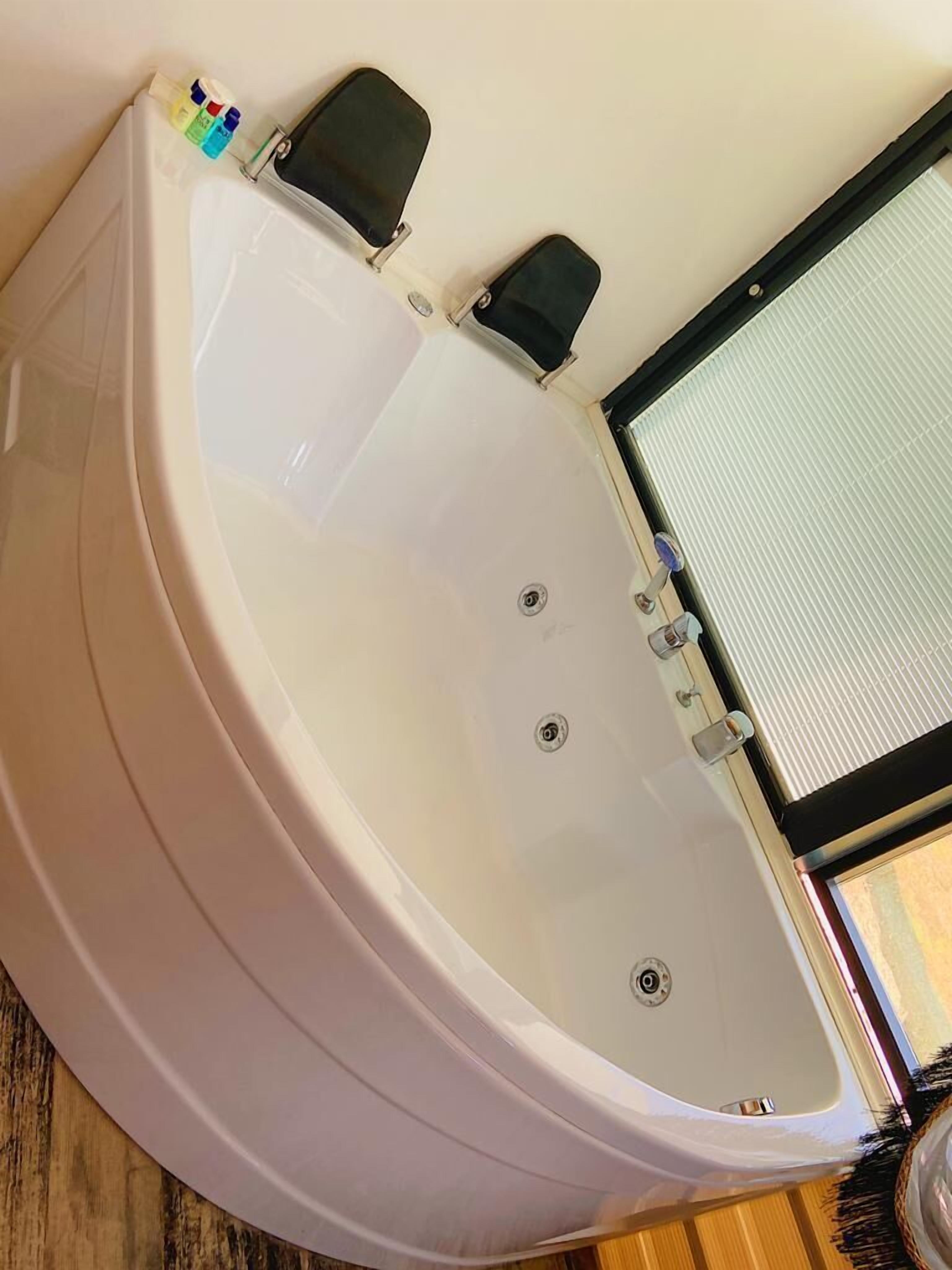 A corner bathtub with a window, a small shelf with toiletries, and a wooden floor.