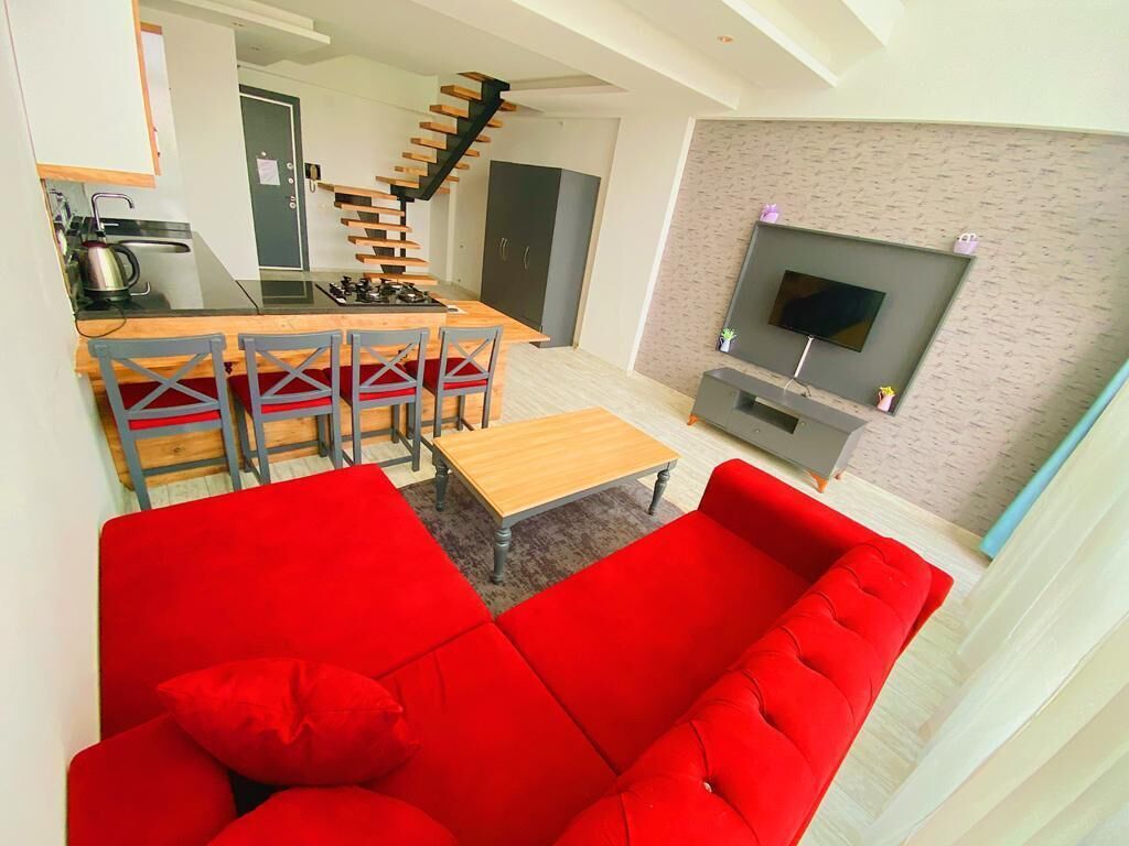 Una sala moderna con un sofá rojo, una mesa de comedor de madera con sillas, una zona de cocina con estufa y un televisor montado en la pared.