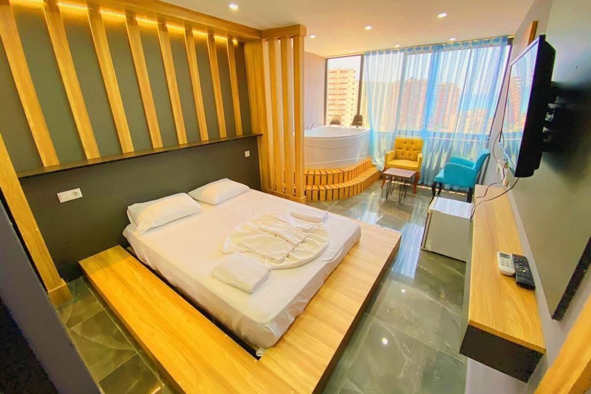 Quarto de hotel moderno com uma cama grande, uma pequena área de estar com uma cadeira e mesa, uma varanda com vista e uma televisão.