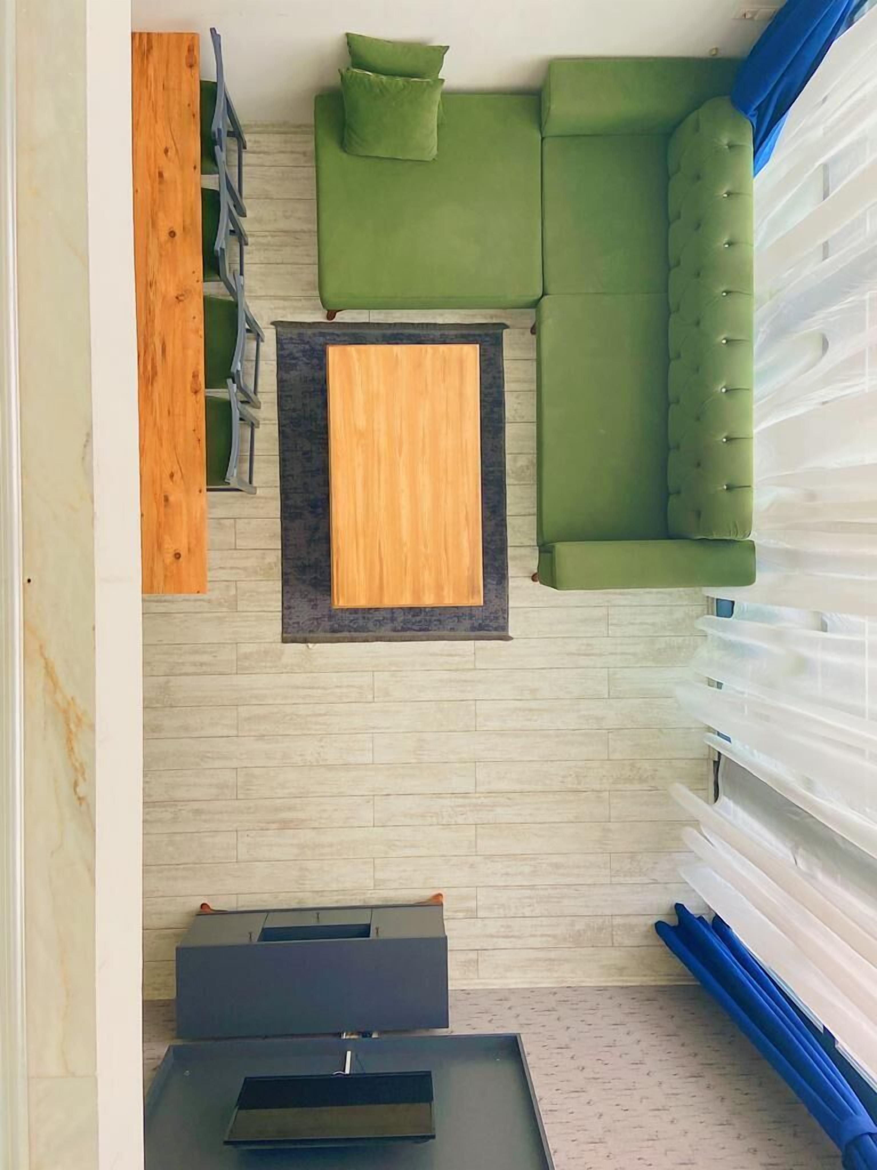 Una habitación con un sofá verde, un aparador de madera, una mesita pequeña y un ventanal con cortinas traslúcidas.