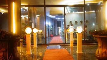 Vestíbulo de hotel con alfombra roja, plantas en macetas grandes y entrada de vidrio.