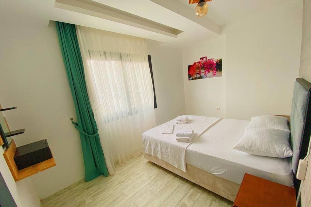 Quarto de hotel com cama, uma mesinha, prateleira com objetos decorativos e janela com cortinas.