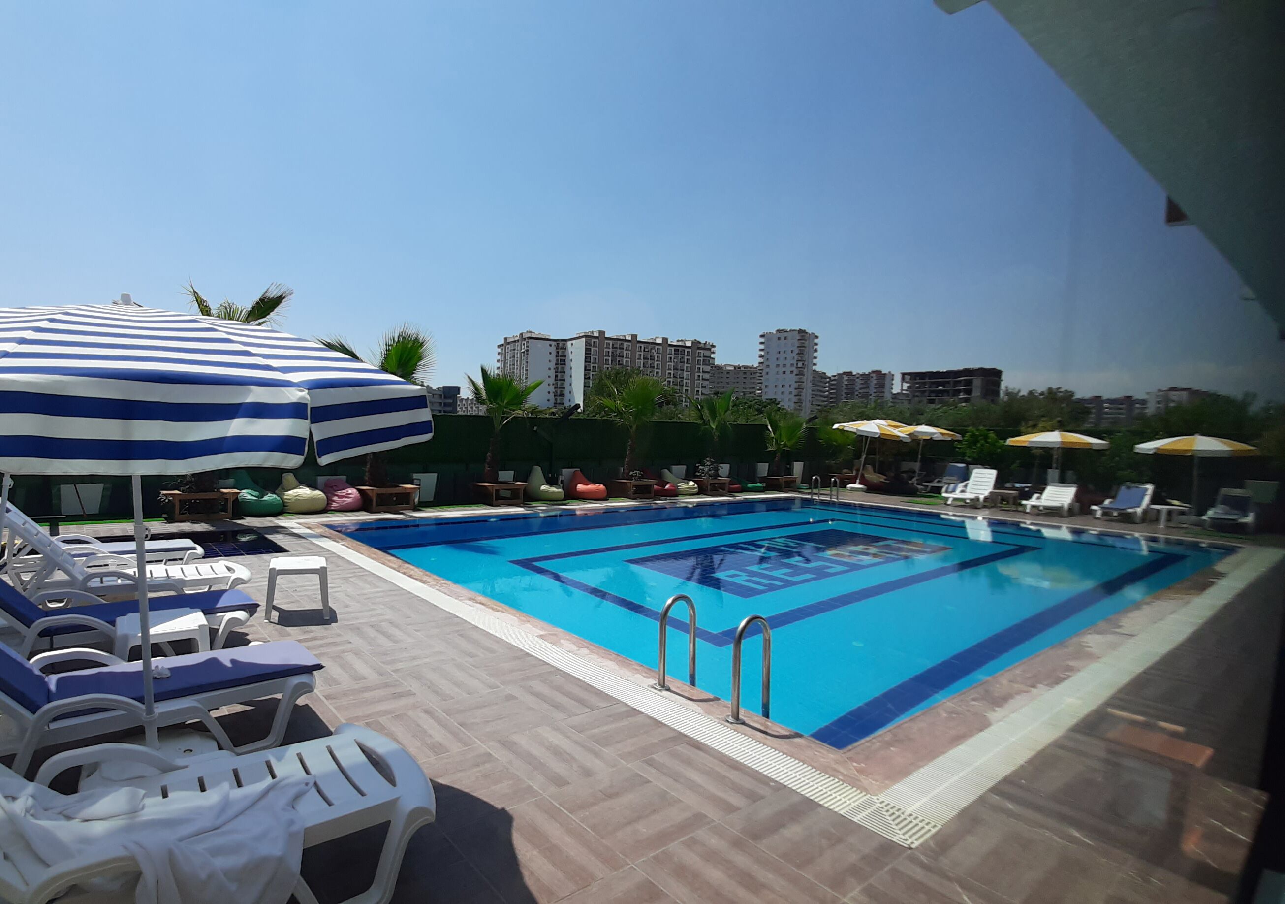 Ein Schwimmbecken mit Liegestühlen und Sonnenschirmen, umgeben von Palmen und Gebäuden im Hintergrund.