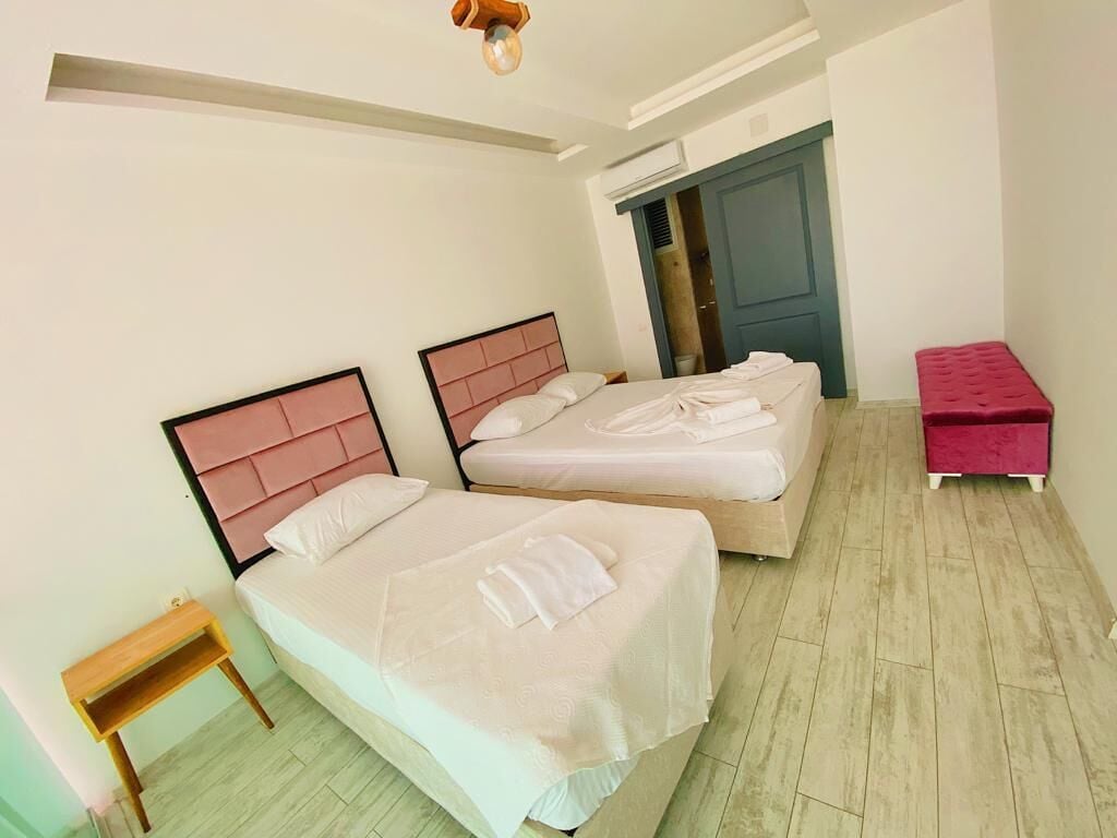 A room with two beds, a small wooden nightstand, a red bench, and a door leading to another room.