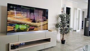 A modern living room with a flat-screen TV mounted on a white cabinet, a potted plant, and a glass coffee table.