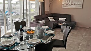 A modern living room with a glass dining table set for four, a dark-colored sofa, and a painting of sailboats on the wall.