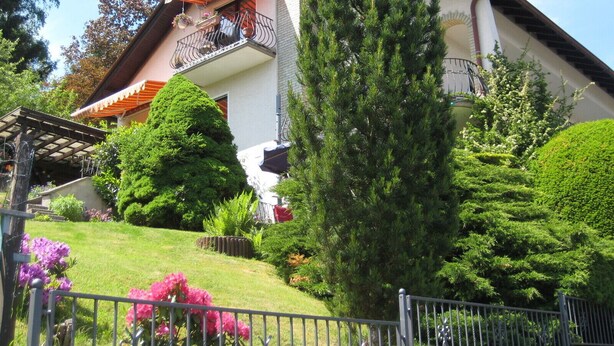 Una casa de dos pisos con balcón, rodeada de jardines y árboles bien cuidados.