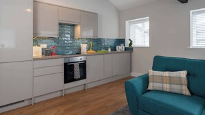 Een moderne keuken met witte kastjes, een inbouwoven en een backsplash van blauwe tegels. Op de voorgrond staat een blauwgroene bank met een geruit kussen.