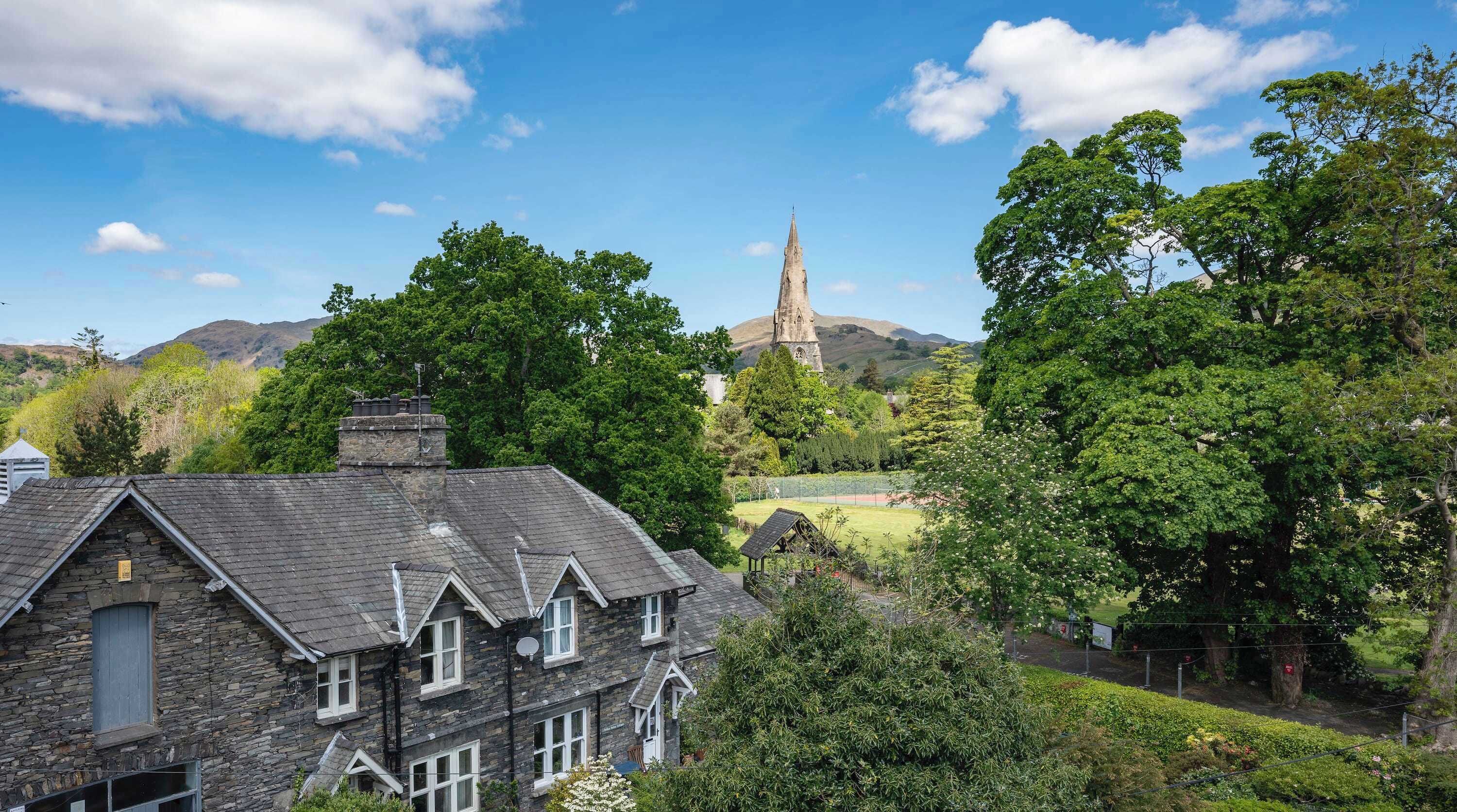 A stone building with a pitched roof, surrounded by lush greenery and a church spire in the background.