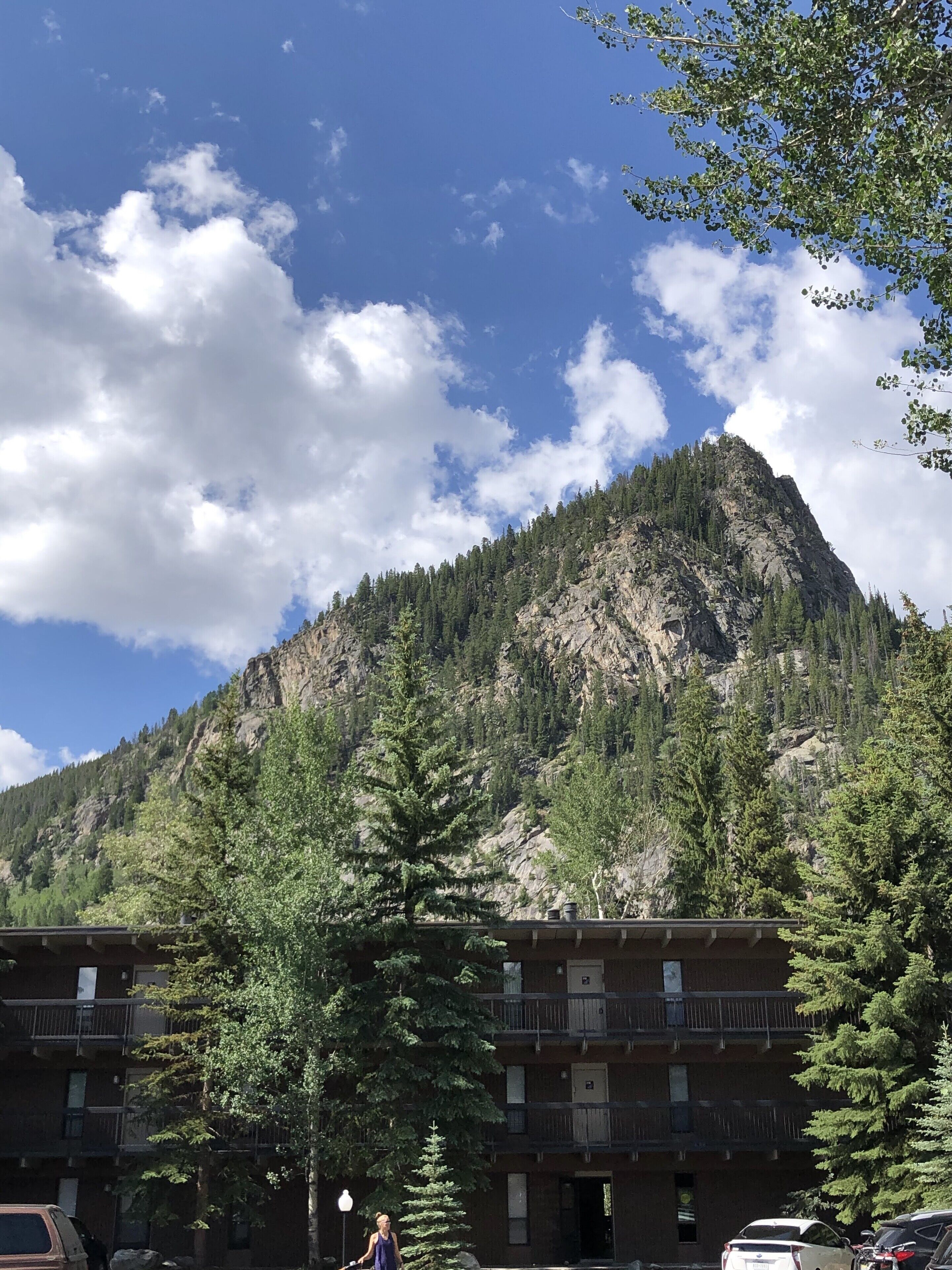 A mountain with a rocky peak and dense forest. There is a building with multiple floors and balconies.