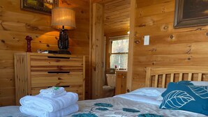 A wooden cabin room with two beds, a wooden dresser, and a window with a view of trees.