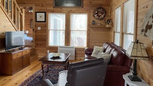 A cozy cabin interior with wooden walls, a ceiling fan, a television, a fireplace, and a large window with blinds.
