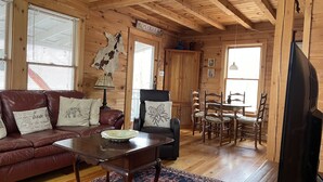 A wooden cabin interior with a leather sofa, a dining table with chairs, a television, and a mounted animal hide.