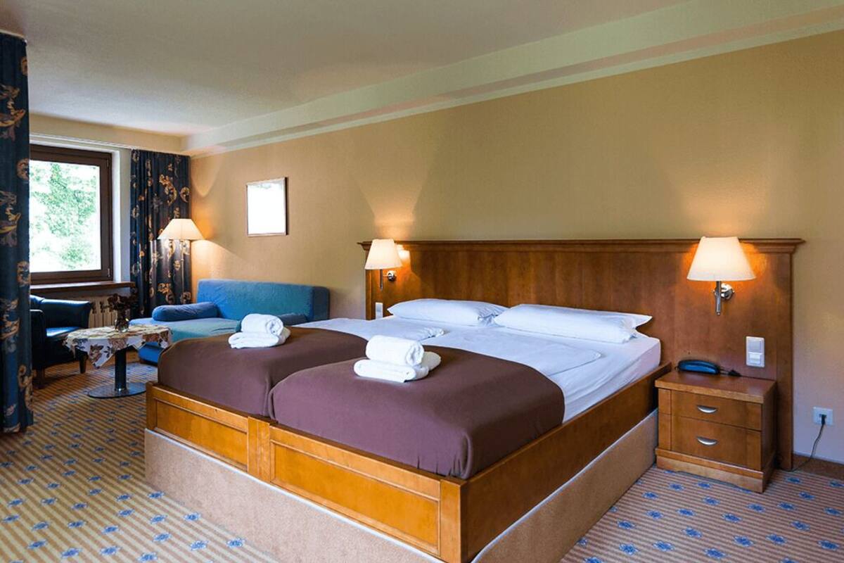 Ein Hotelzimmer mit einem Doppelbett, zwei Nachttischen, einem kleinen Schreibtisch mit Stuhl, einem Sofa und einem Fenster mit Vorhängen.