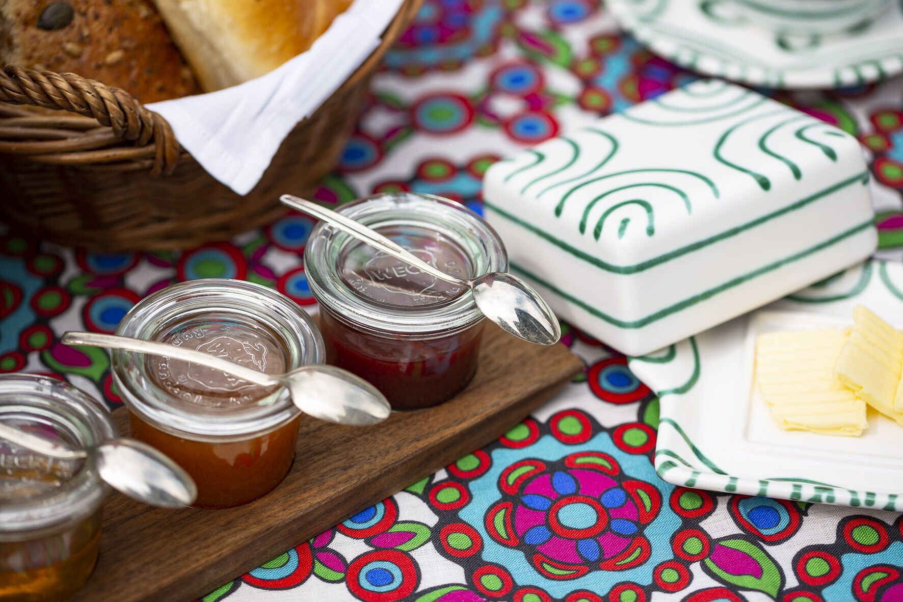 Jars of honey and butter on a wooden board, with a colorful tablecloth and a basket of bread in the background.