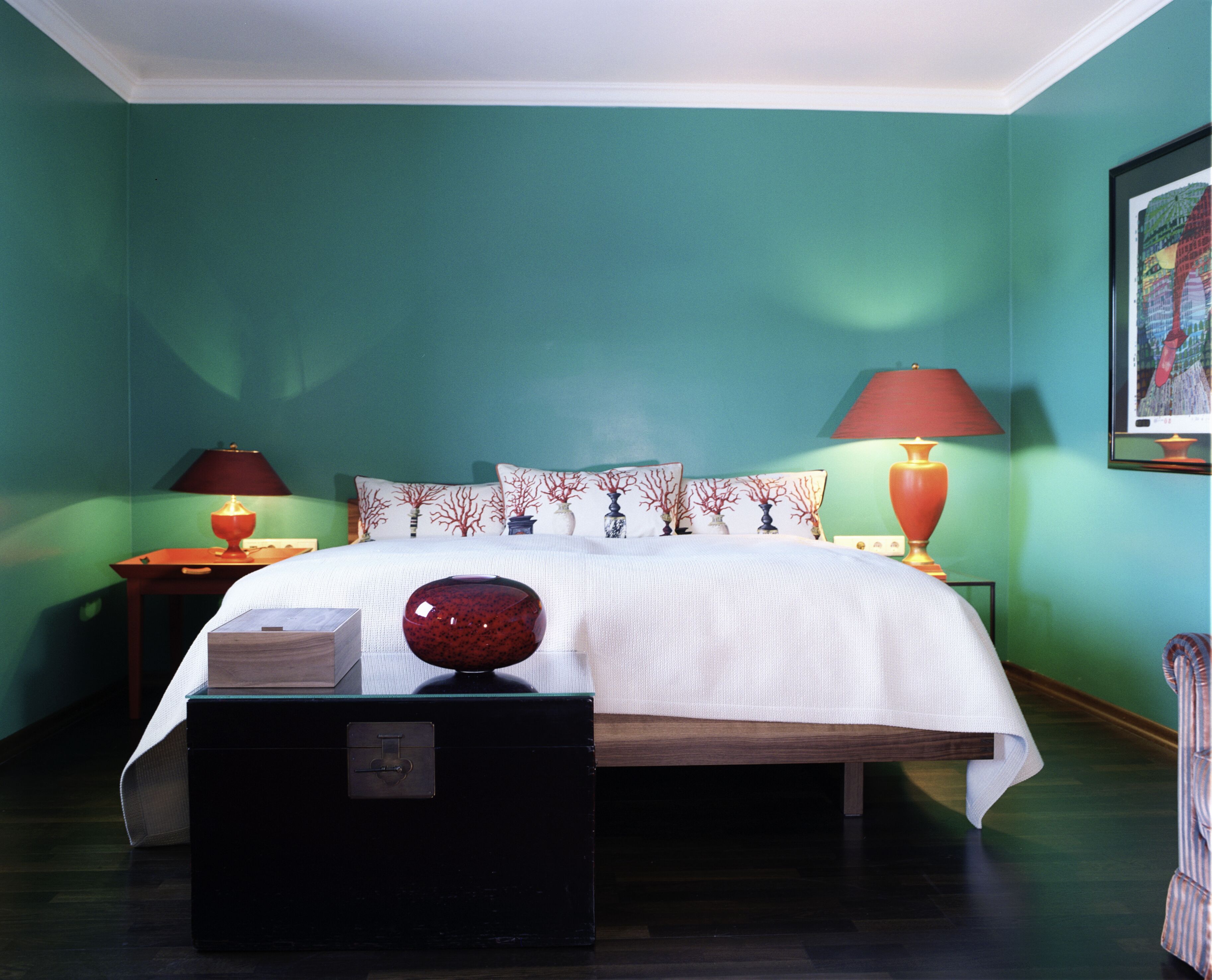 A neatly made bed with a white comforter and decorative pillows, two bedside lamps, a small wooden nightstand with a lamp, and a black storage box.