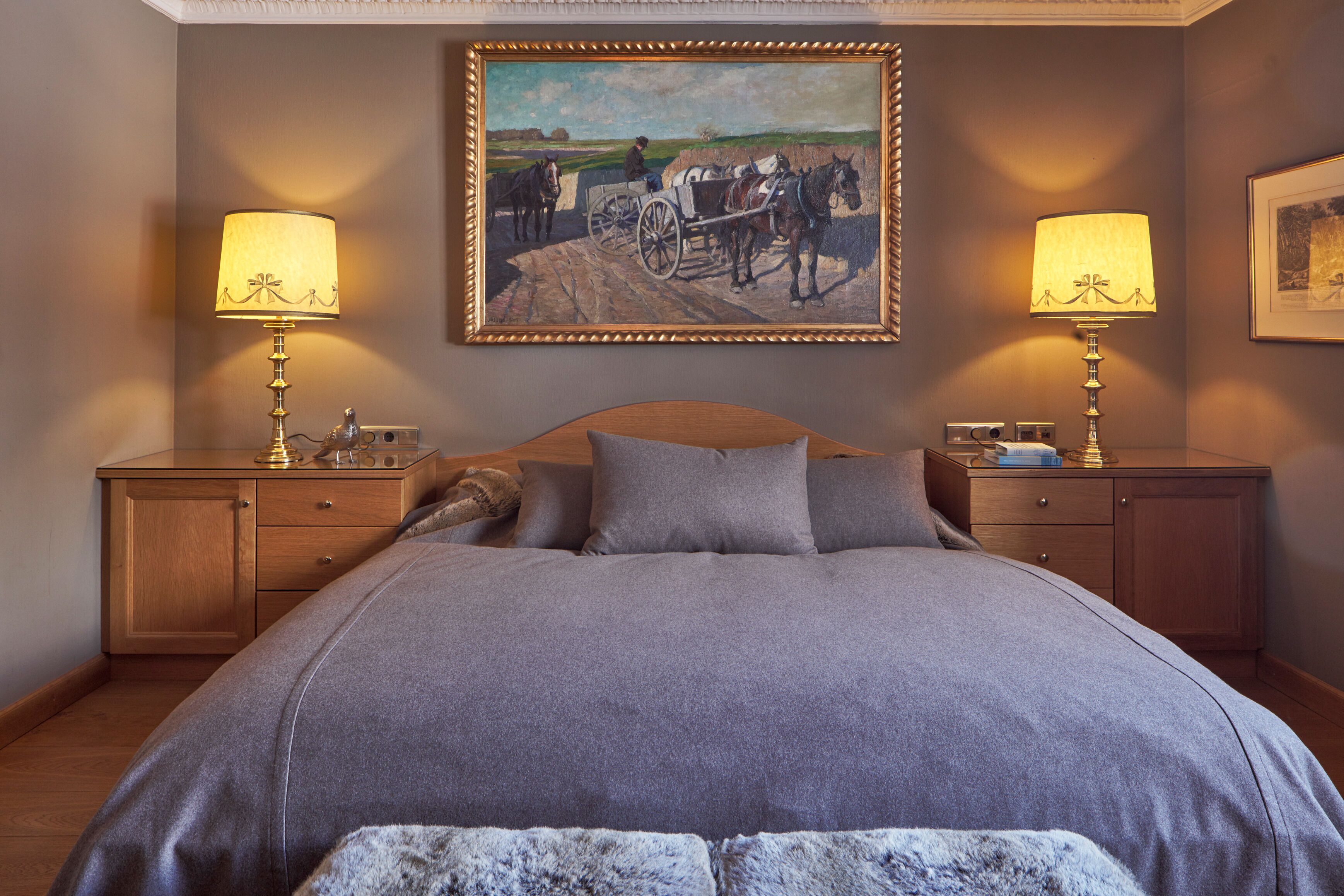 A bedroom with a large bed, two bedside tables, a nightstand, a wooden headboard, a painting above the bed, and two lamps on the bedside tables.